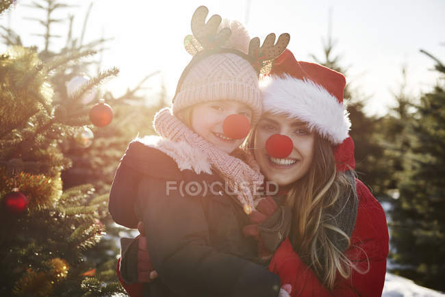 Niña y madre en el bosque de árboles de Navidad con narices rojas, retrato - foto de stock