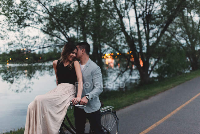 Romántico joven susurrando a su novia en el manillar de bicicleta por el lago al atardecer - foto de stock