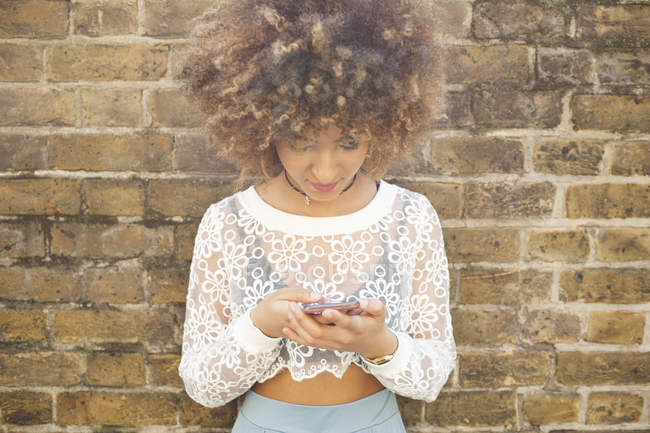 Portrait of Young smiling woman using smartphone against brick wall — Stock Photo
