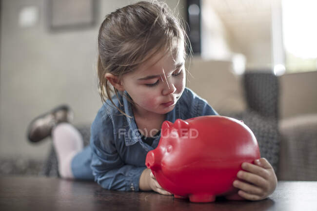 Menina deitada na mesa de café olhando para o banco piggy vermelho — Fotografia de Stock
