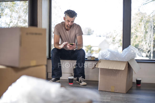 Uomo seduto in casa non arredata circondata da scatole di cartone e utilizzando smartphone — Foto stock