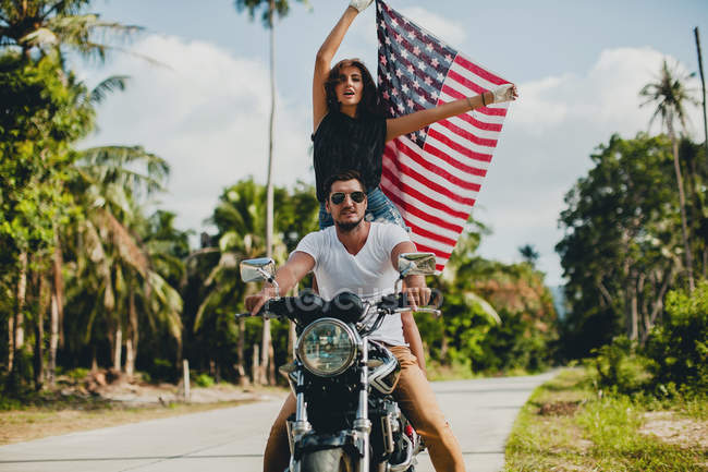 Coppia giovane che sorregge la bandiera americana mentre guida una moto su strada rurale, Krabi, Thailandia — Foto stock
