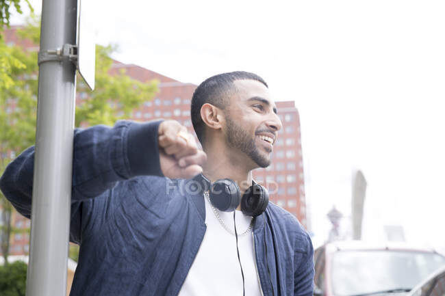 Young man leaning against lamppost, headphones around neck, smiling — Stock Photo