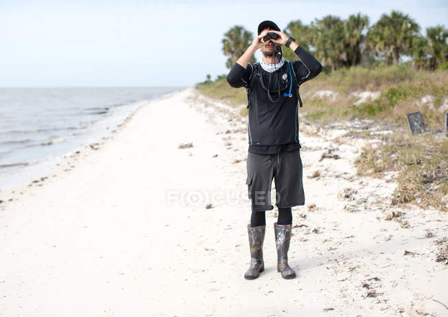 Vista frontal del hombre mirando a través de los prismáticos cuando está de pie en la playa - foto de stock