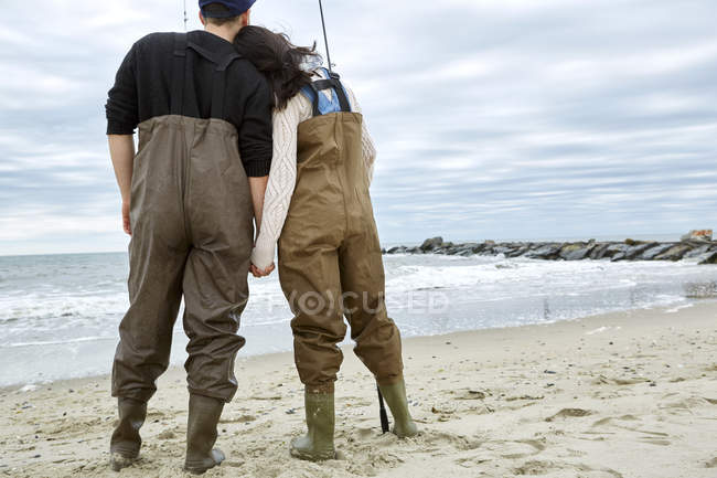 Pareja joven en vadeadores de pesca en la playa - foto de stock