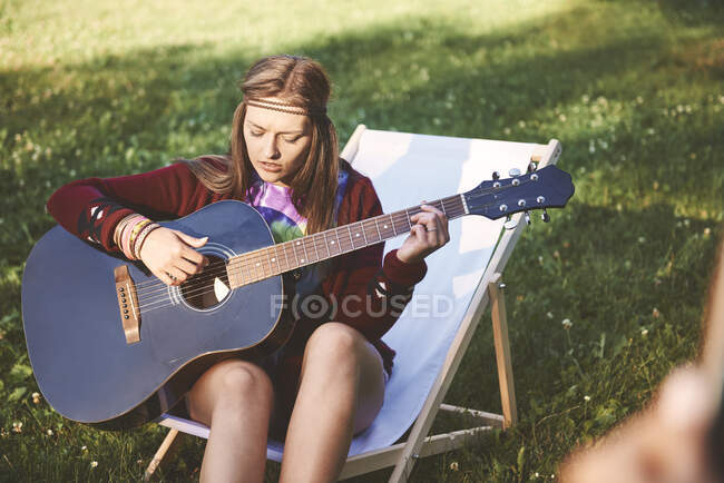 Jovem boho mulher sentada na cadeira de praia tocando guitarra acústica no festival — Fotografia de Stock