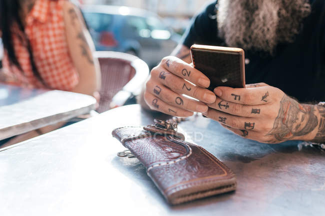 Зрілий хіпстер використовує смартфон у тротуарному кафе, крупним планом татуйовану руку — стокове фото