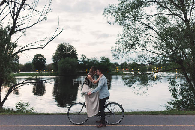 Romântico jovem casal na bicicleta cara a cara por lago ao entardecer — Fotografia de Stock
