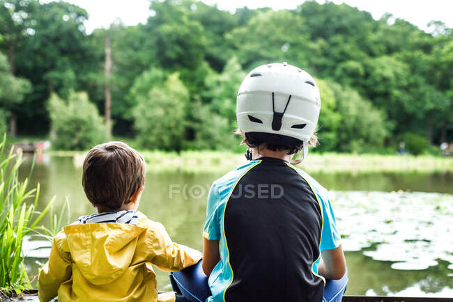Due giovani fratelli seduti sul bordo dell'acqua, fratello maggiore con casco da ciclismo, vista posteriore — Foto stock