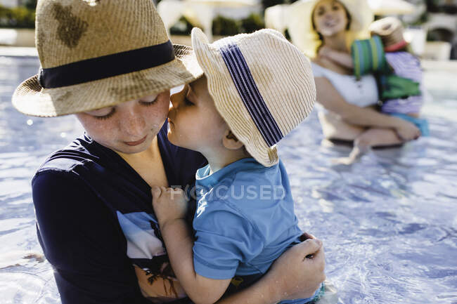 Family in outdoor swimming pool, young boy holding younger brother — Stock Photo