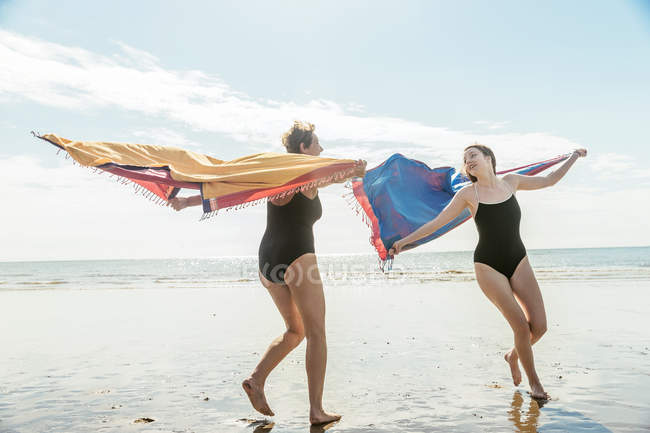 Мать и дочь бегут по пляжу с шалями в воздухе — стоковое фото