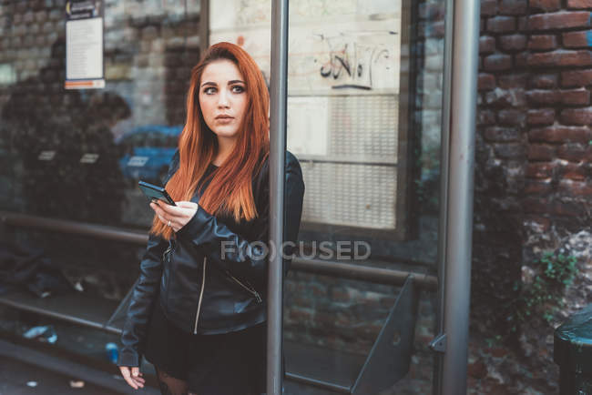 Red haired woman at bus stop holding smartphone — Stock Photo