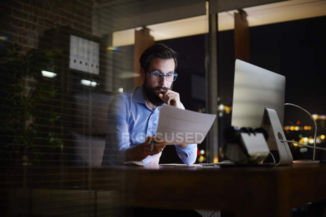 Young businessman reading paperwork at office desk at night — Stock Photo