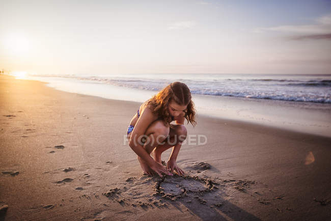 Girl drawing heart in sand on beach — Stock Photo