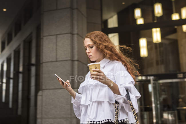 Mujer joven con café para llevar mirando el teléfono inteligente en la acera, Nueva York, Estados Unidos - foto de stock