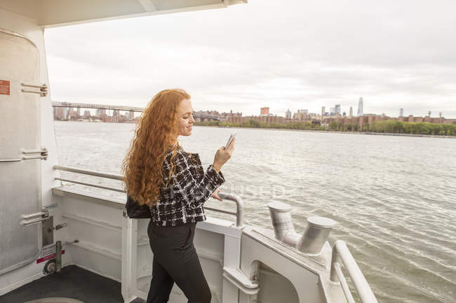 Молодая деловая женщина на палубе паром глядя на смартфон, Нью-Йорк, США — стоковое фото
