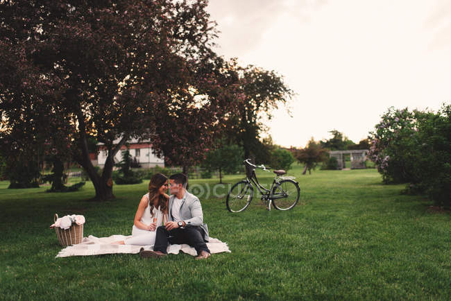 Romántica pareja joven tomando un picnic de champán rosa en el parque al atardecer - foto de stock