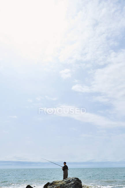 Distant view of young man standing on rock sea fishing — Stock Photo