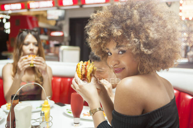 Três jovens amigos comendo hambúrgueres sentados no restaurante — Fotografia de Stock