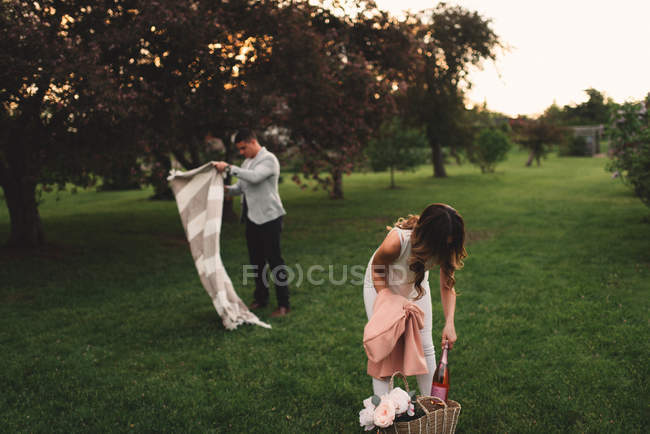 Casal jovem preparando cobertor de piquenique e champanhe rosa no parque ao entardecer — Fotografia de Stock