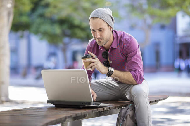 Joven al aire libre, sentado en el banco, usando el ordenador portátil, sosteniendo la taza de café para llevar - foto de stock