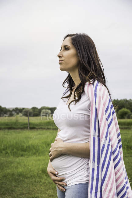 Giovane donna incinta in piedi in campo, tenendo lo stomaco, coperta intorno alle spalle, espressione pensierosa — Foto stock