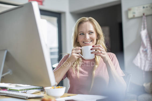 Woman at computer drinking coffee and smiling — Stock Photo