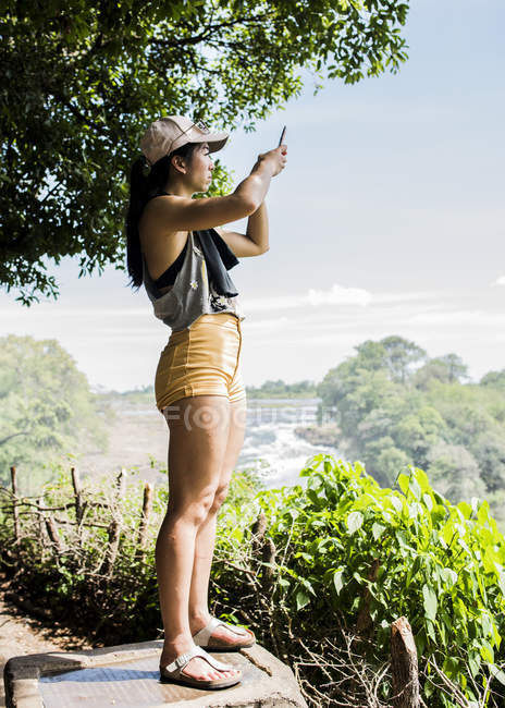 Seitenansicht einer jungen Touristin, die Fotos mit dem Smartphone von Victoria-Wasserfällen macht, Zimbabwe, Afrika — Stockfoto