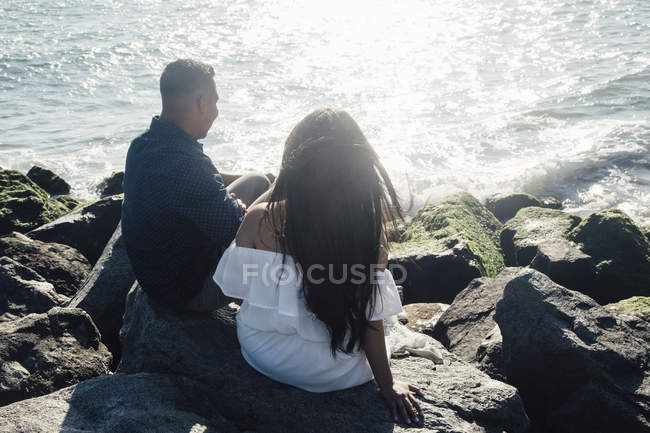 Pareja sentada sobre rocas costeras, mirando a la vista, vista trasera - foto de stock