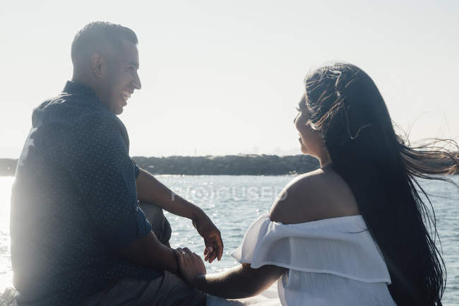 Пара сидящих на прибрежных скалах, держащихся за руки, улыбающихся, с видом сзади — стоковое фото