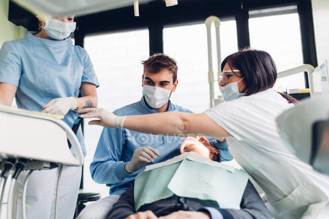 Dentista mirando en la boca del paciente masculino, enfermeras dentales preparando equipo - foto de stock