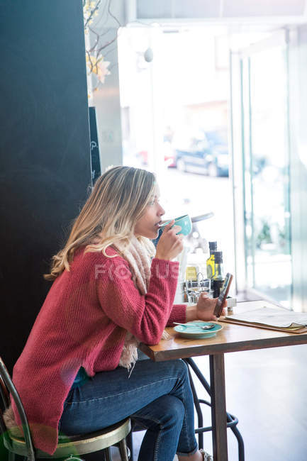Mujer bebiendo café y sosteniendo teléfono inteligente en la cafetería - foto de stock