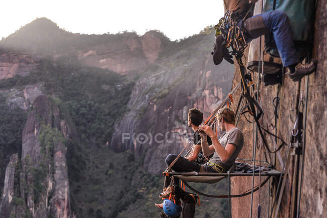 Dos escaladores sentados en portaledge, mirando a la vista, Liming, provincia de Yunnan, China - foto de stock