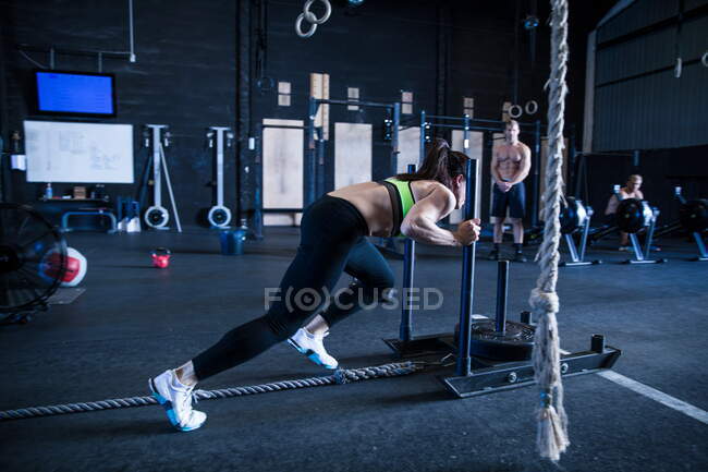 Mujer haciendo ejercicio en el gimnasio, entrenamiento en trineo, amigos en el fondo, viendo - foto de stock
