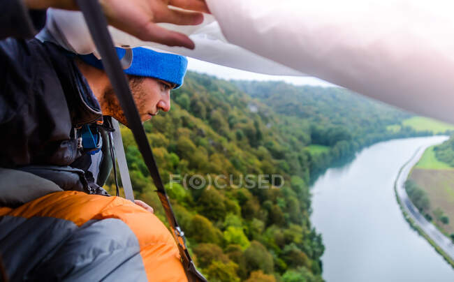 Joven escalador de roca macho mirando desde el portaledge de la cara de roca mirando hacia el río Mosa, Freyr, Bélgica, vista elevada - foto de stock