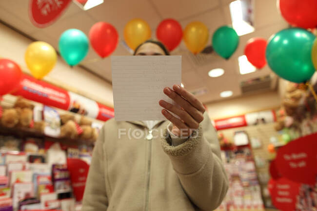Adolescente na loja com lista de compras cobrindo rosto — Fotografia de Stock