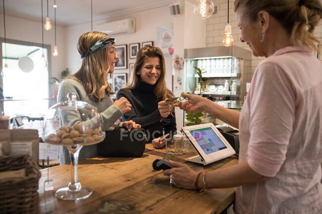 Dos amigas, de pie en el mostrador en la cafetería, entregando la tarjeta de crédito al asistente de la tienda para el pago - foto de stock