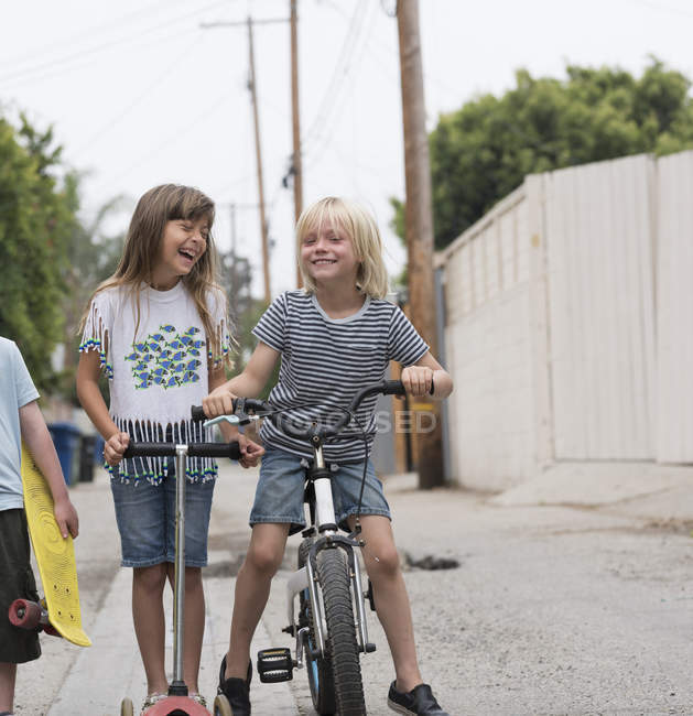 Fille et garçons dans la voie avec scooter, vélo et planche à roulettes — Photo de stock