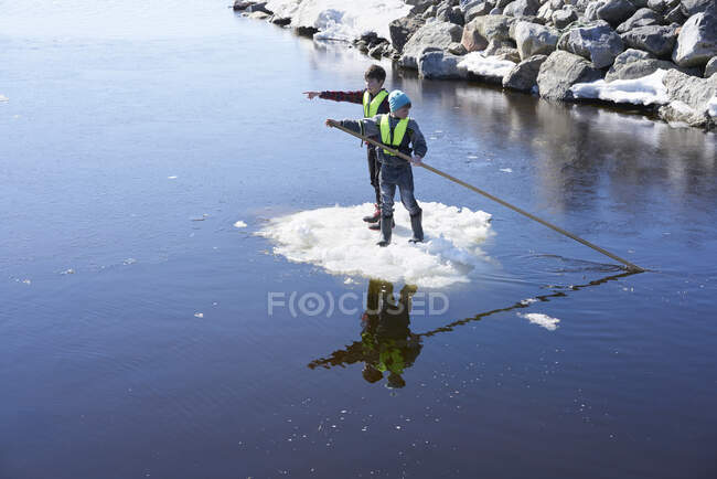 Dos chicos parados en el hielo, en el lago, empujándose junto con el poste - foto de stock