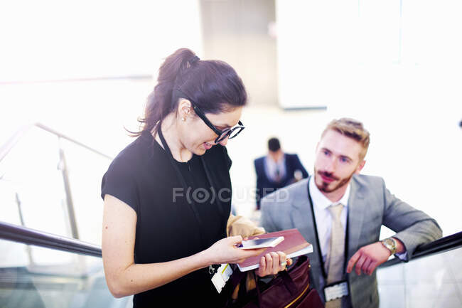 Jovem empresária e homem na escada rolante do aeroporto olhando para o smartphone — Fotografia de Stock
