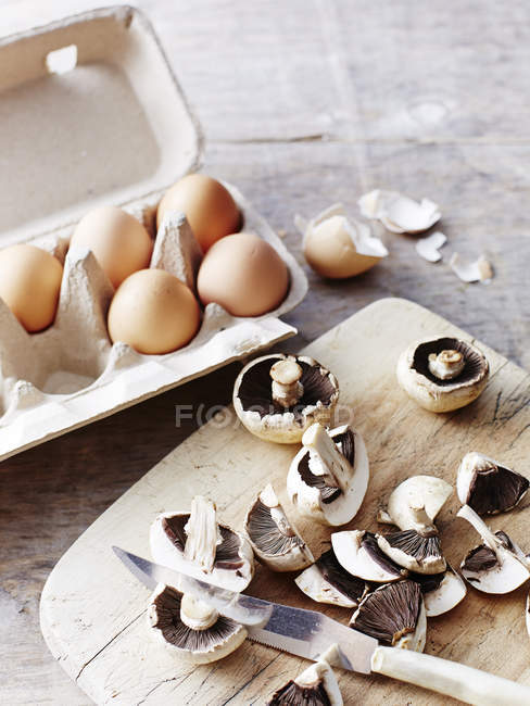 Close-up view of fresh raw mushrooms on cutting board with knife and eggs in egg box — Stock Photo