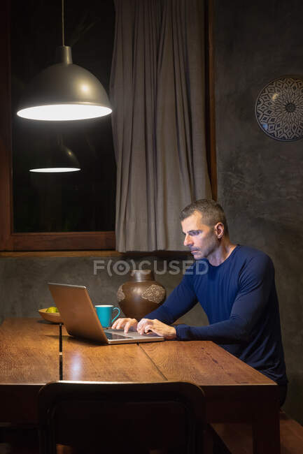 Зрілий чоловік за обіднім столом друкує на ноутбуці, ніч — стокове фото