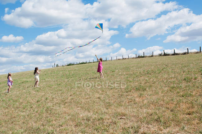 Three girls playing with flying kite in field — Stock Photo