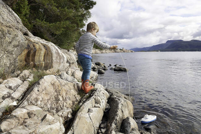 Boy standing on rock by fjord playing with toy boat, Aure, More og Romsdal, Noruega - foto de stock