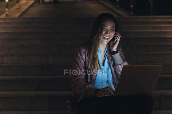 Mujer sentada en escalones y usando laptop con smartphone - foto de stock