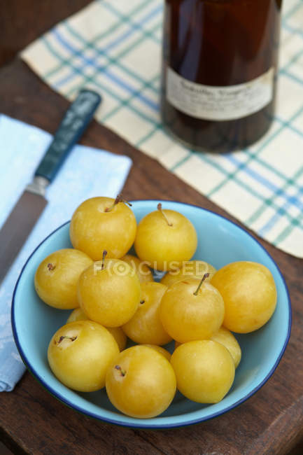 Tigela de ameixas amarelas frescas, garrafa e faca na mesa na cozinha — Fotografia de Stock