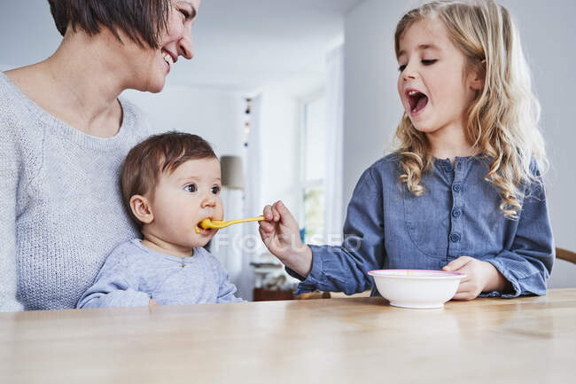 Familia sentada en la mesa de la cocina, joven niña cuchara-alimentación hermanita - foto de stock