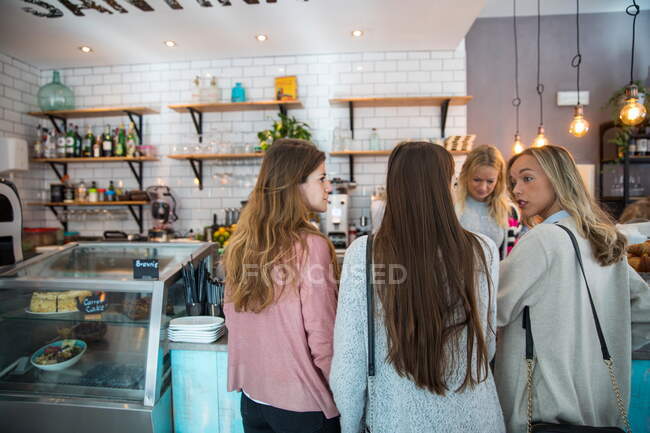 Tres amigas, de pie en el mostrador en la cafetería, vista trasera - foto de stock