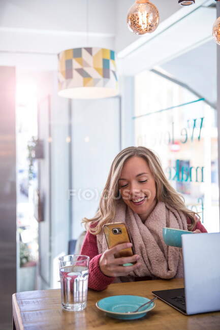Donna che usando smartphone e tazza di caffè della holding della seduta in caffè — Foto stock