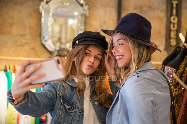 Due amici in negozio, provando cappelli, scattando selfie, usando smartphone — Foto stock